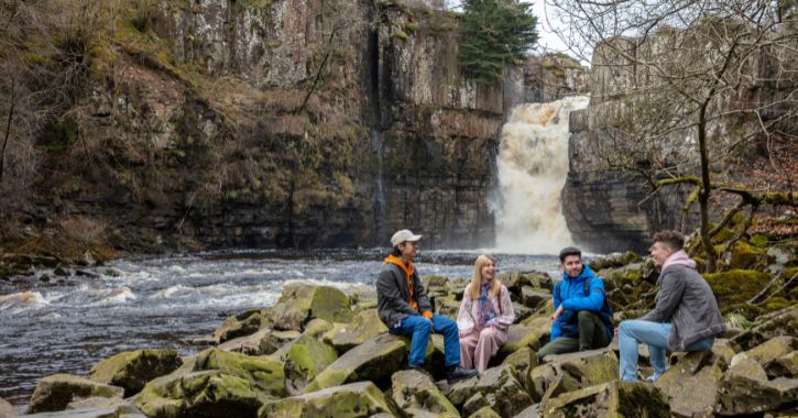 group of young people sat on rocks smiling and laughing with High Force Waterfall in background.